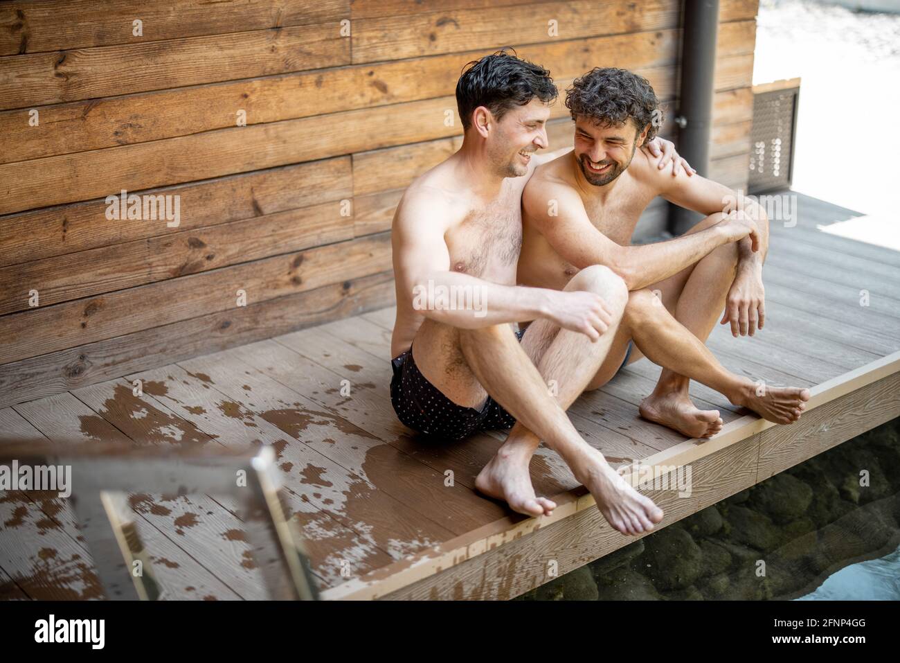 Amici maschi seduti sulla terrazza vicino al lago nel complesso termale e parlando. Rilassatevi e divertitevi nel centro benessere. Abbracci e ridendo insieme. Foto Stock