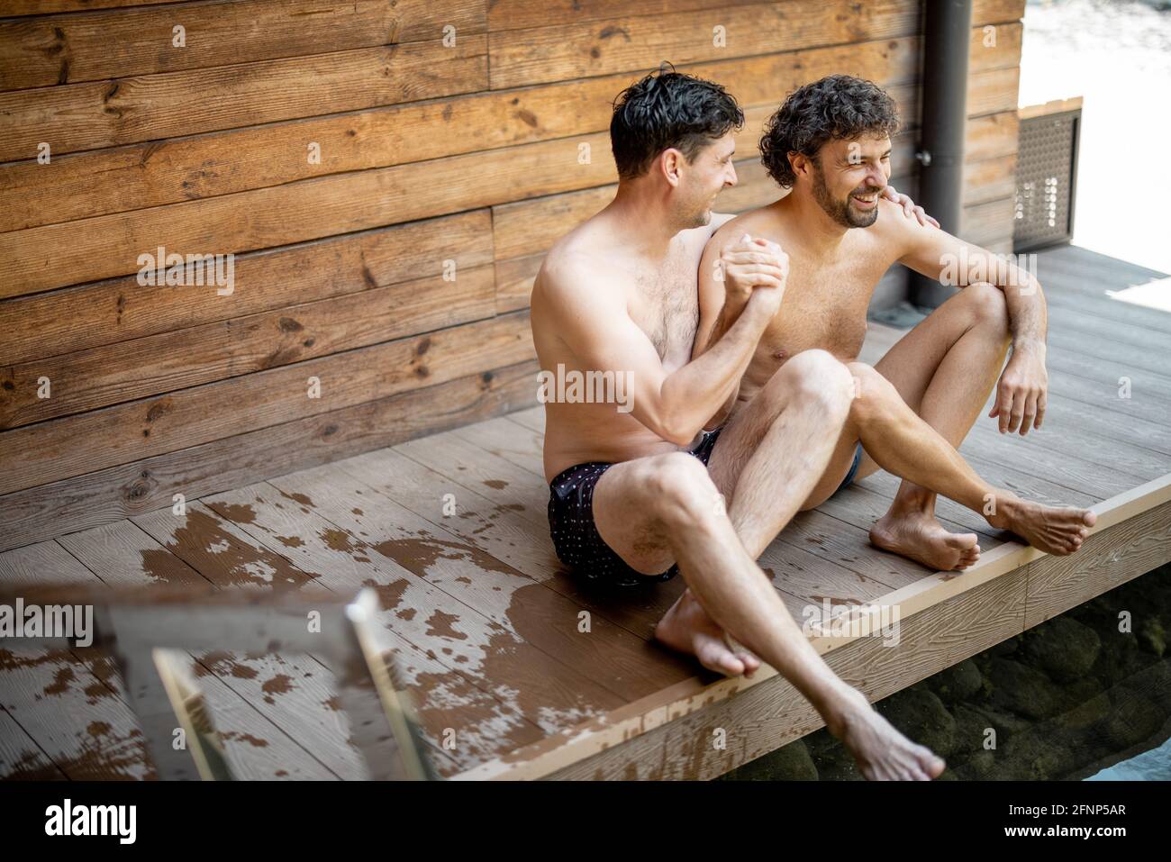 Amici maschi seduti sulla terrazza vicino al lago nel complesso termale mentre parlano e tengono le mani. Rilassatevi e divertitevi nel centro benessere. Foto Stock