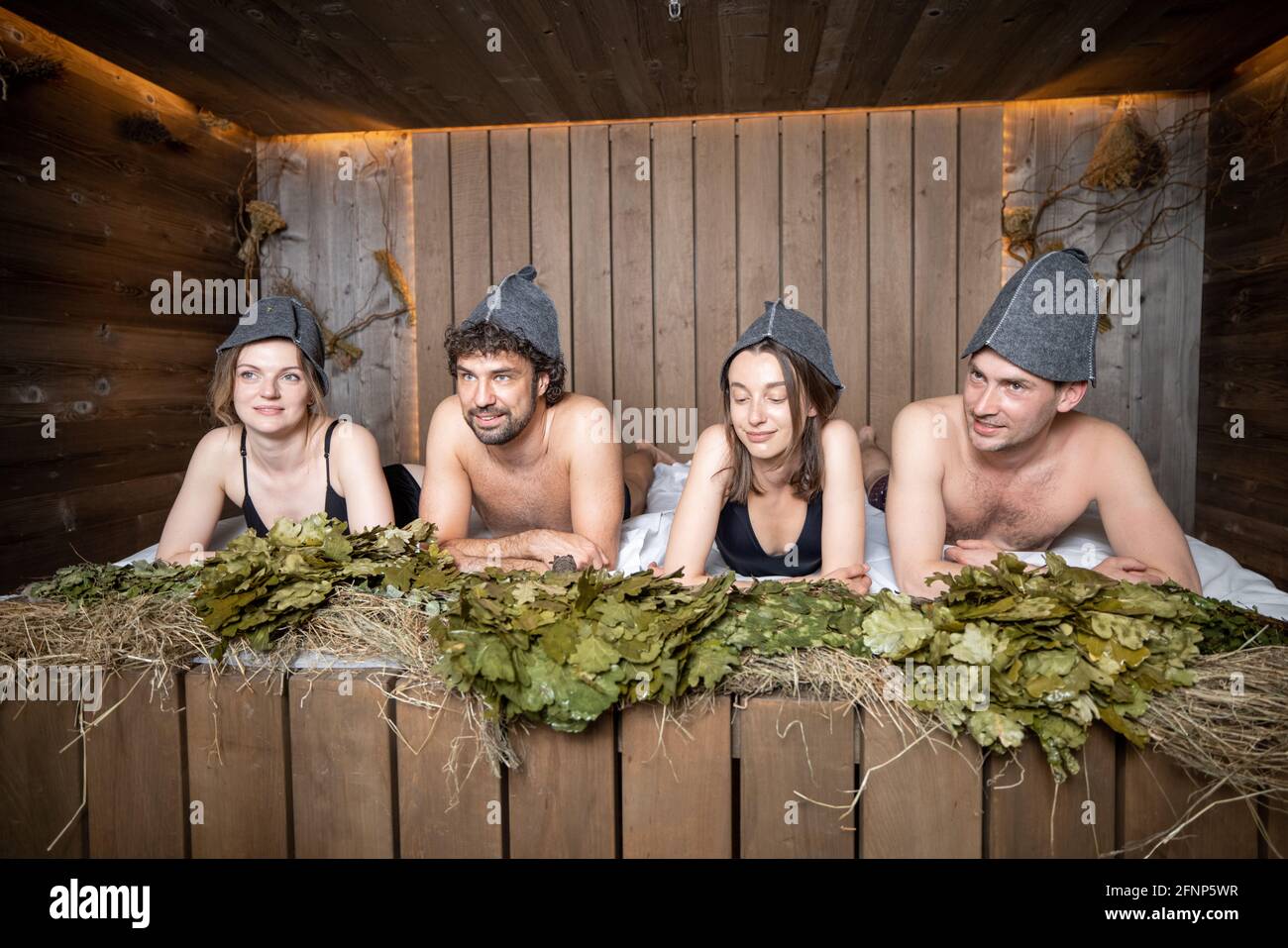 Compagnia di amici sdraiati su una panca di legno coperta di fieno nella sauna russa tradizionale in cappelli sauna. Benessere e svago in spa. Foto Stock