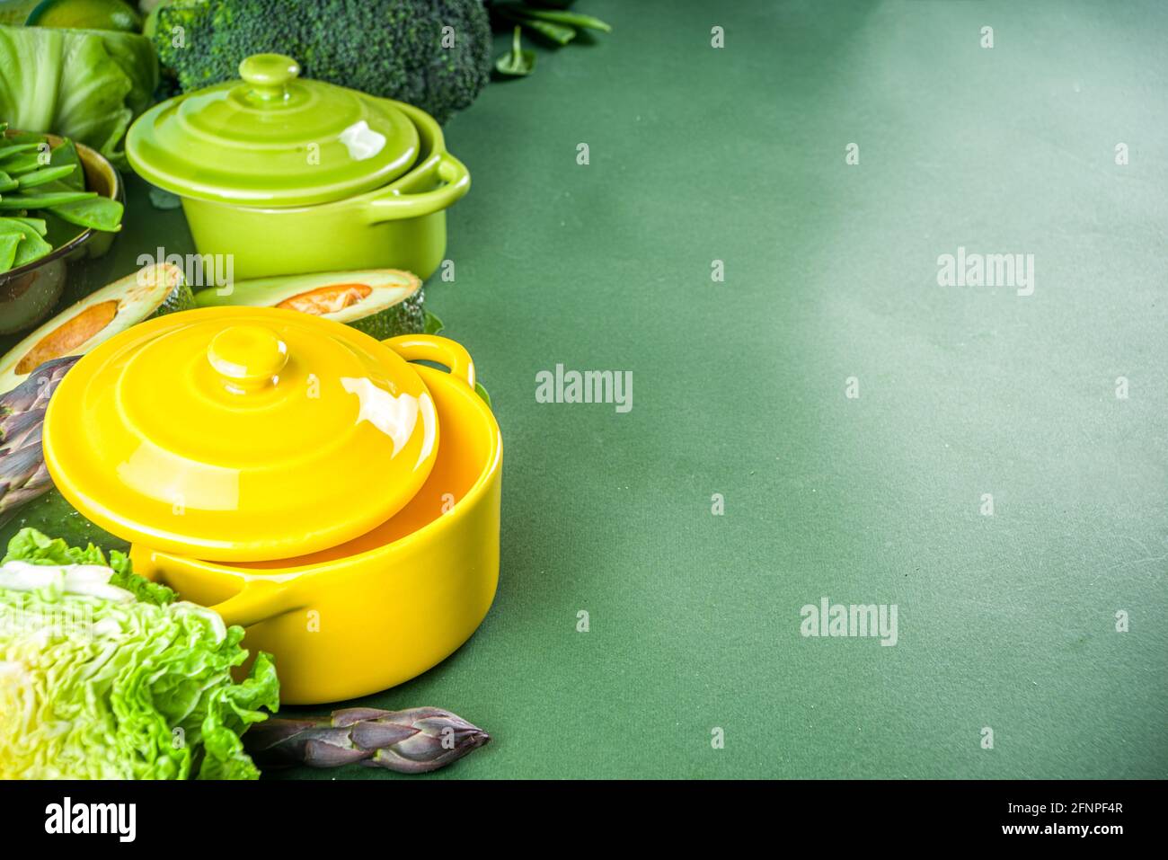 Vari ingredienti vegetali di primavera verdi organici con pentole da cucina vuote e colorate su un tavolo da cucina verde scuro, vista dall'alto. Dieta zuppa sana Foto Stock