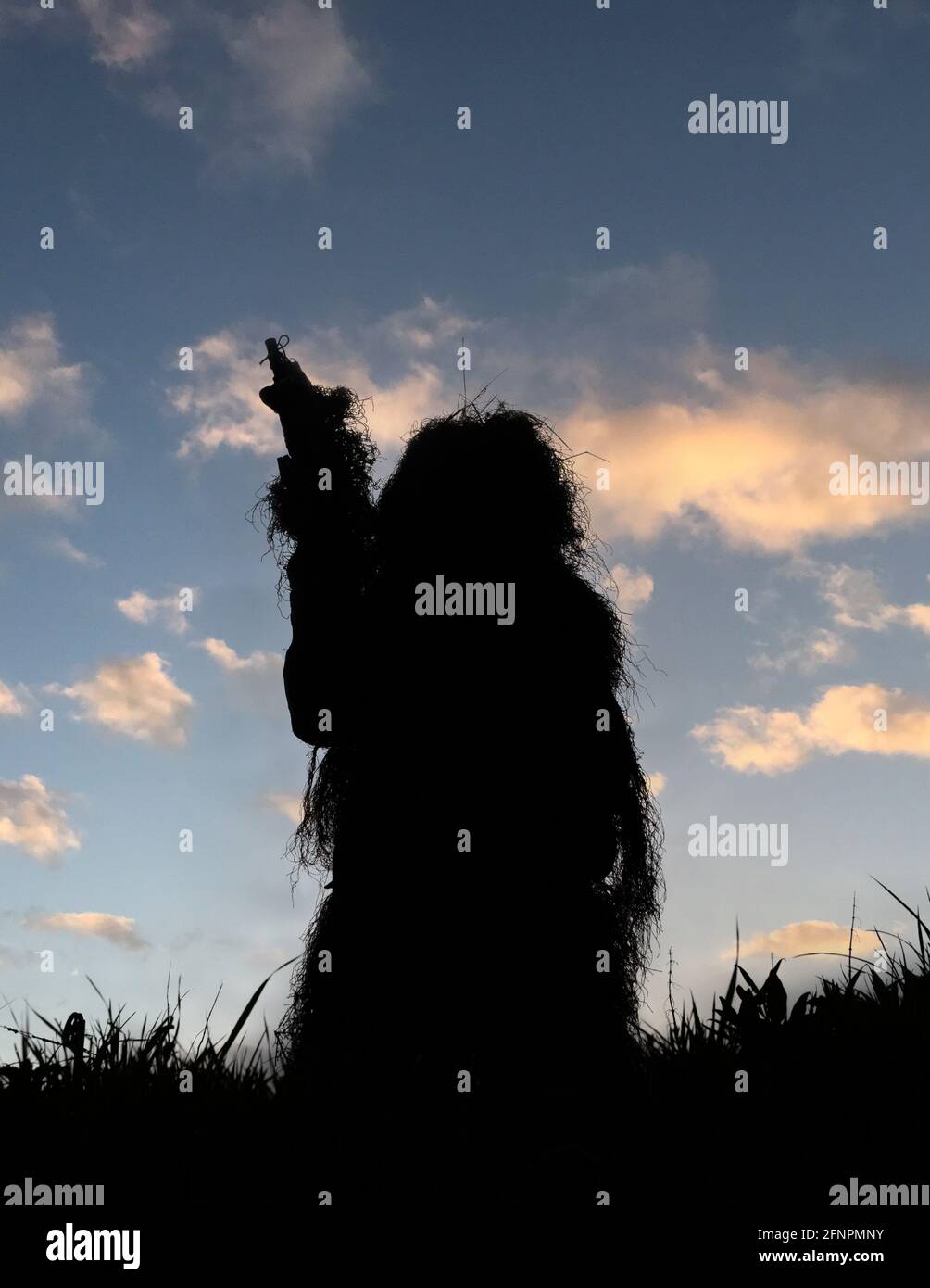 Sniper Marksman in camouflage completo Ghillie con silhouette contro A. Blue Sky in piedi sulla cima di una collina che guarda sopra Il campo di battaglia dopo una vittoria Foto Stock