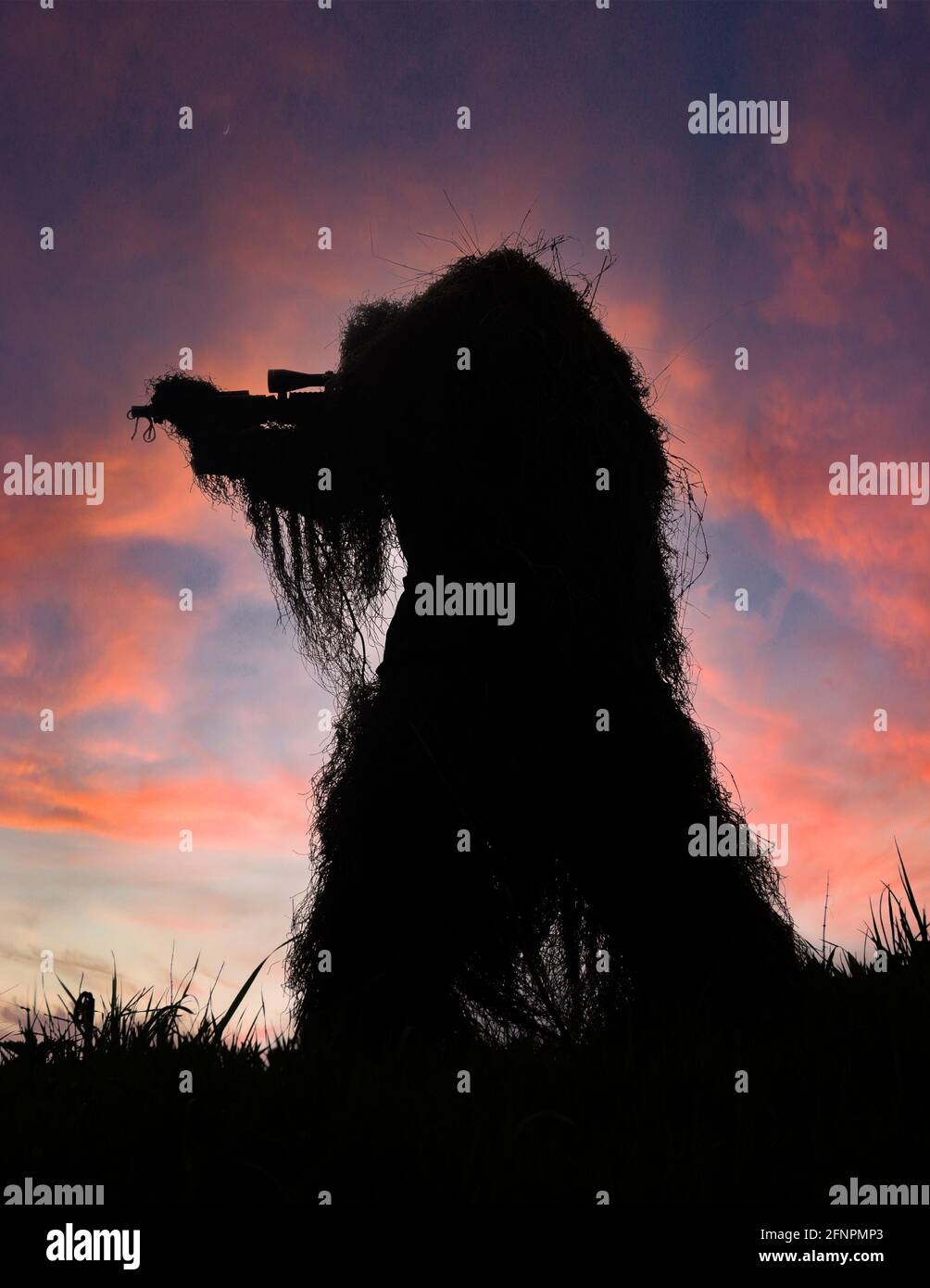 Paintball Sniper in Full Ghillie Suit Camouflage si è fatto strada contro un Sunset Sky sulla cima di una collina con la sua arma disegnata, puntata e pronta a sparare Foto Stock