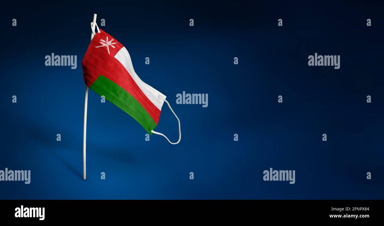 Maschera Oman su sfondo blu scuro. Bandiera ondulata di Oman dipinta su maschera medica sul palo. Flag di attacco del virus. Concetto della bandiera della lotta contro Foto Stock