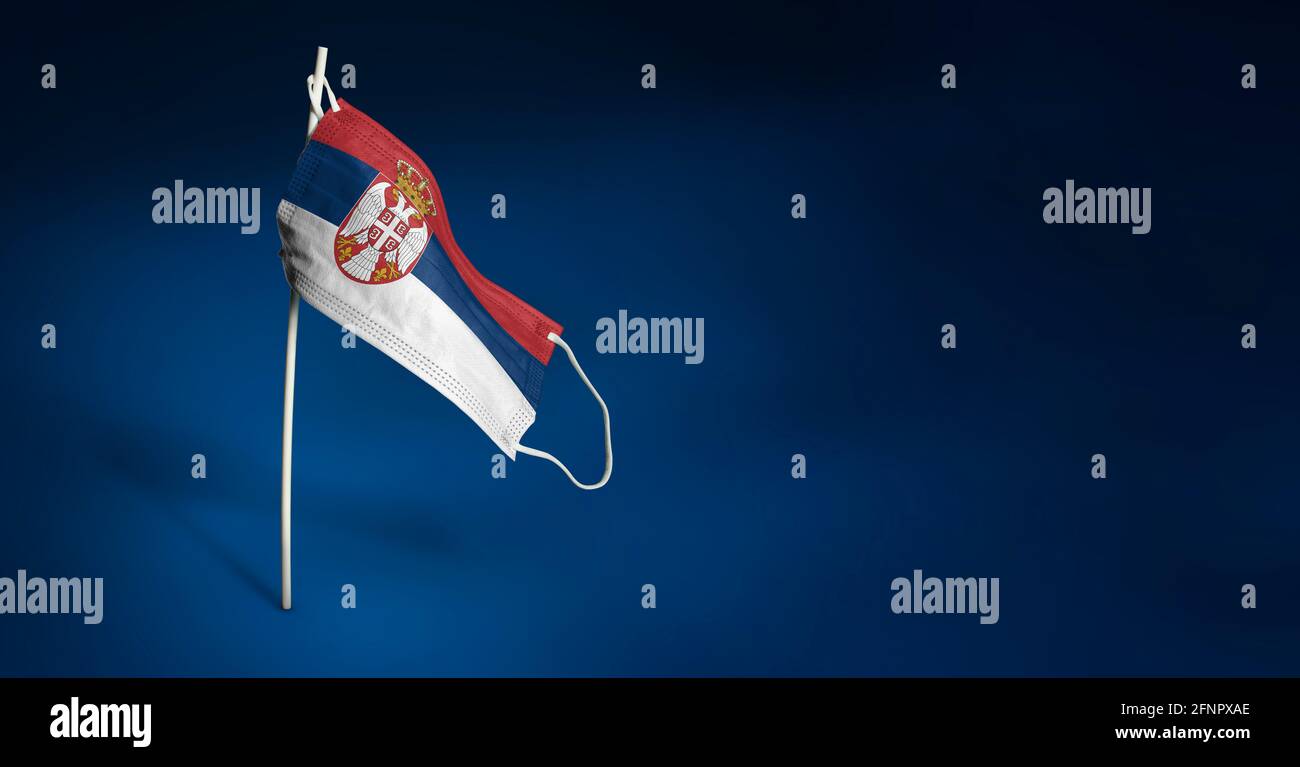 Maschera Serbia su sfondo blu scuro. Bandiera ondulata della Serbia dipinta su maschera medica sul palo. Flag di attacco del virus. Concetto della bandiera della lotta agai Foto Stock
