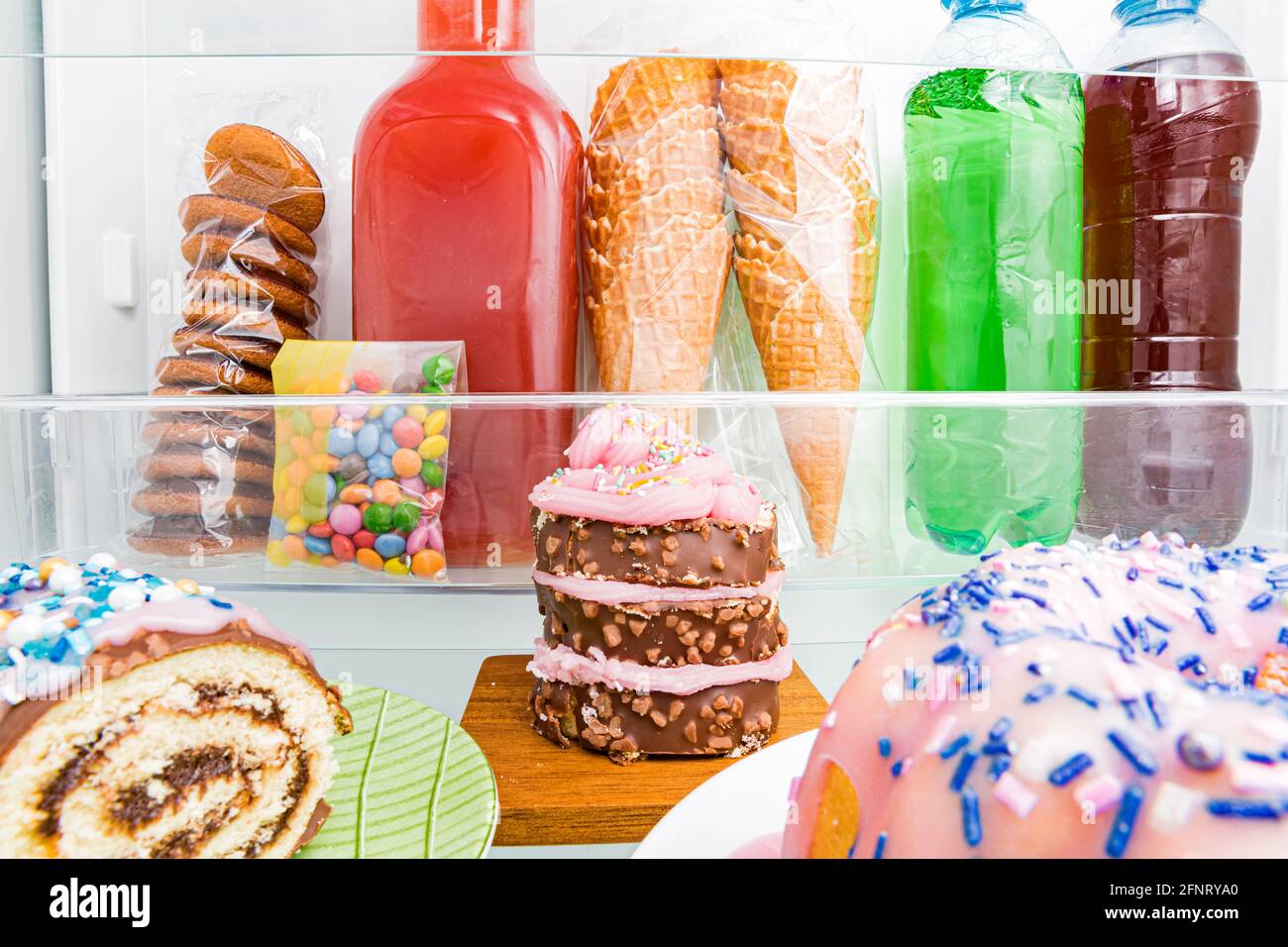Primo piano frigorifero dall'interno, ripiani in vetro con dolci colorati, torte, biscotti, caramelle, bottiglie di bevande zuccherate. Alimentazione malsana, zucchero cibo con Foto Stock