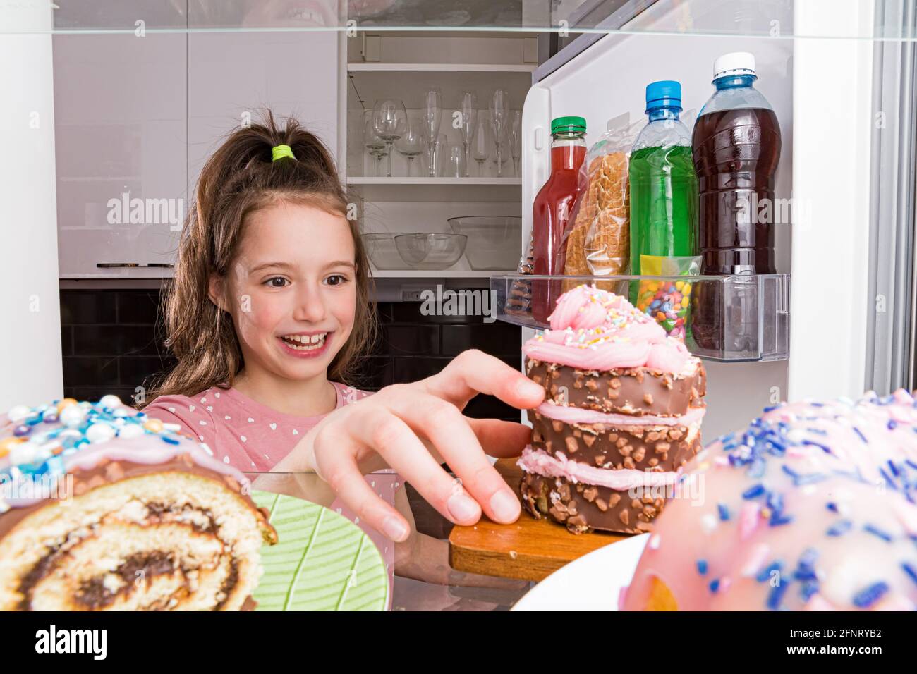 Frigorifero aperto dall'interno, ripiani in vetro con dolci colorati, torte, biscotti, caramelle, bottiglie di zucchero. Alimentazione malsana, cibo da zucchero Foto Stock