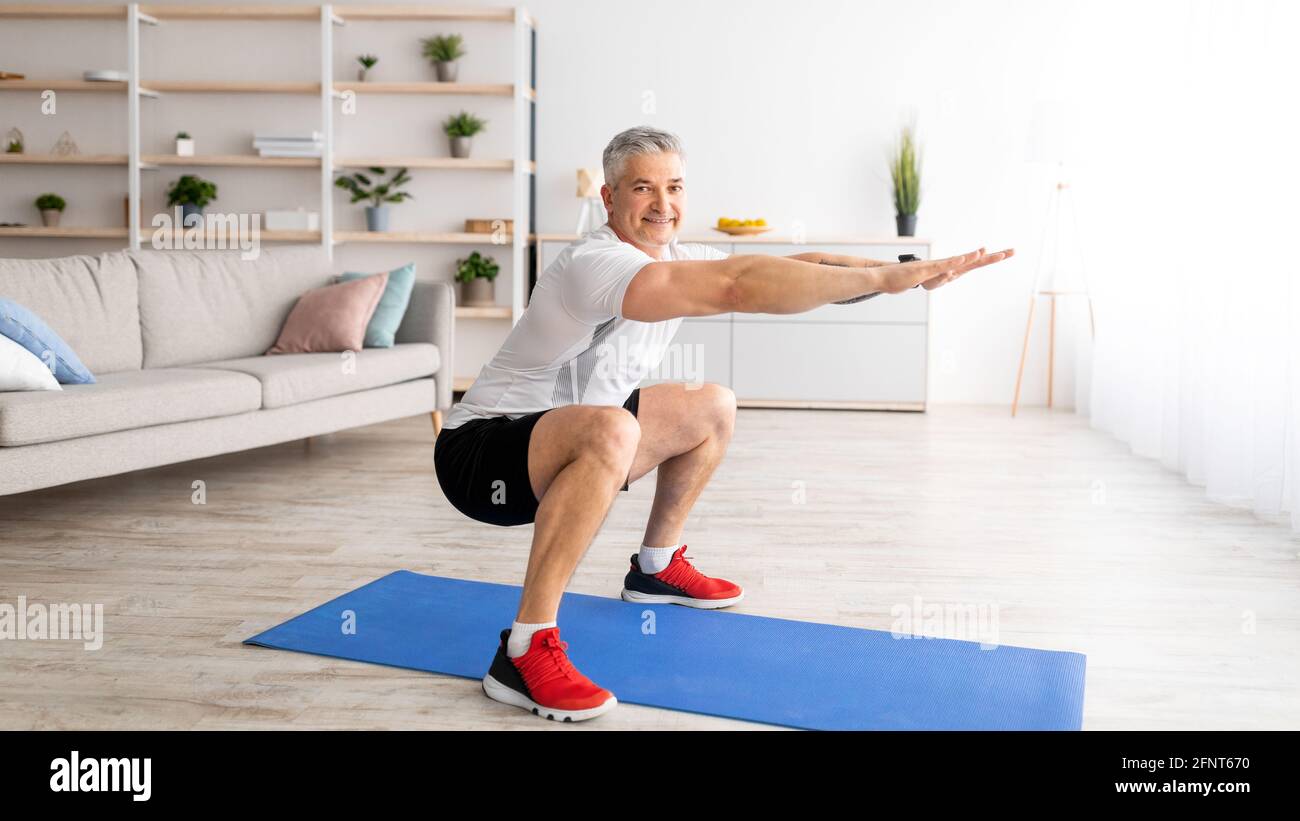 Concetto di allenamento domestico. Uomo anziano felice in sportswear che fa squats all'interno del soggiorno Foto Stock