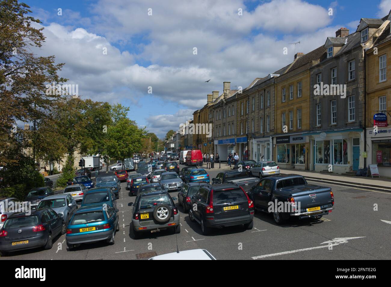 Chipping Norton centro città in Oxfordshire, Regno Unito con case, negozi e auto parcheggiate in una giornata di sole. La piazza si inclina leggermente a sinistra Foto Stock