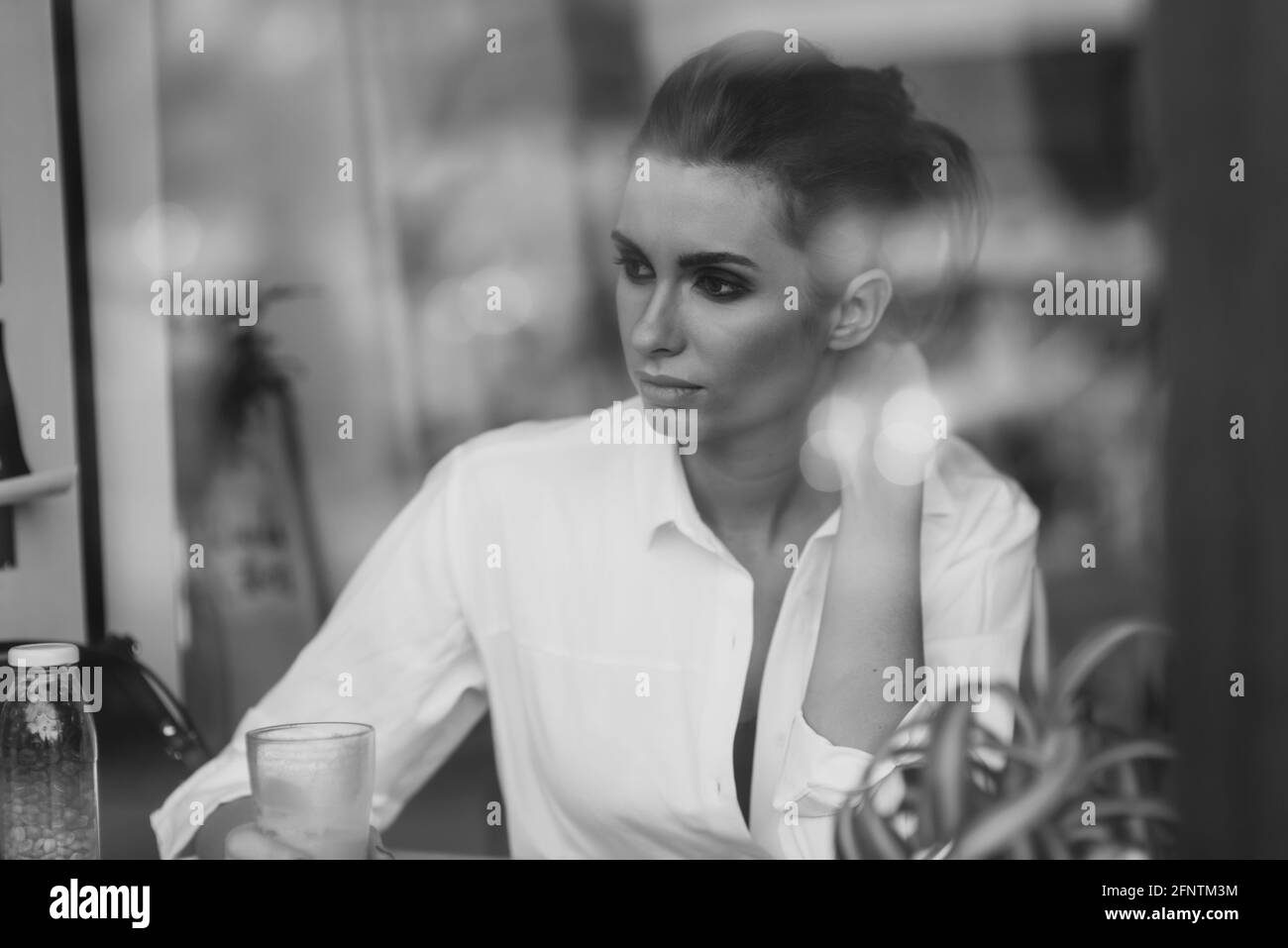 Una bella giovane ragazza si siede in un caffè dietro un bicchiere con una tazza di caffè e guarda attentamente fuori dalla finestra. Foto d'arte in bianco e nero tramite gla Foto Stock