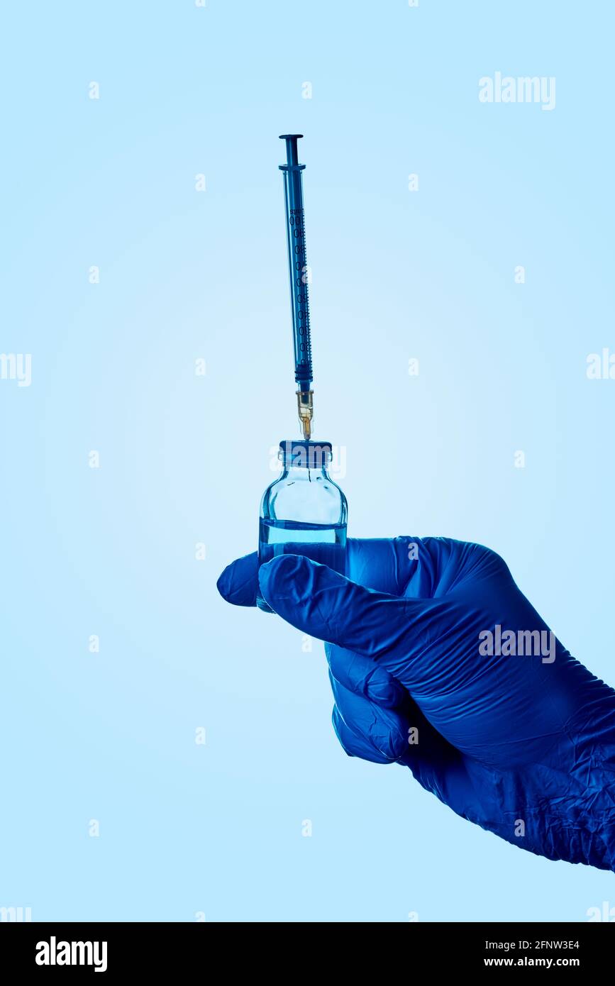 chiusura di un operatore sanitario o di laboratorio, indossando guanti chirurgici blu, tenendo un flaconcino con un liquido blu e una siringa bloccata nel suo tappo Foto Stock