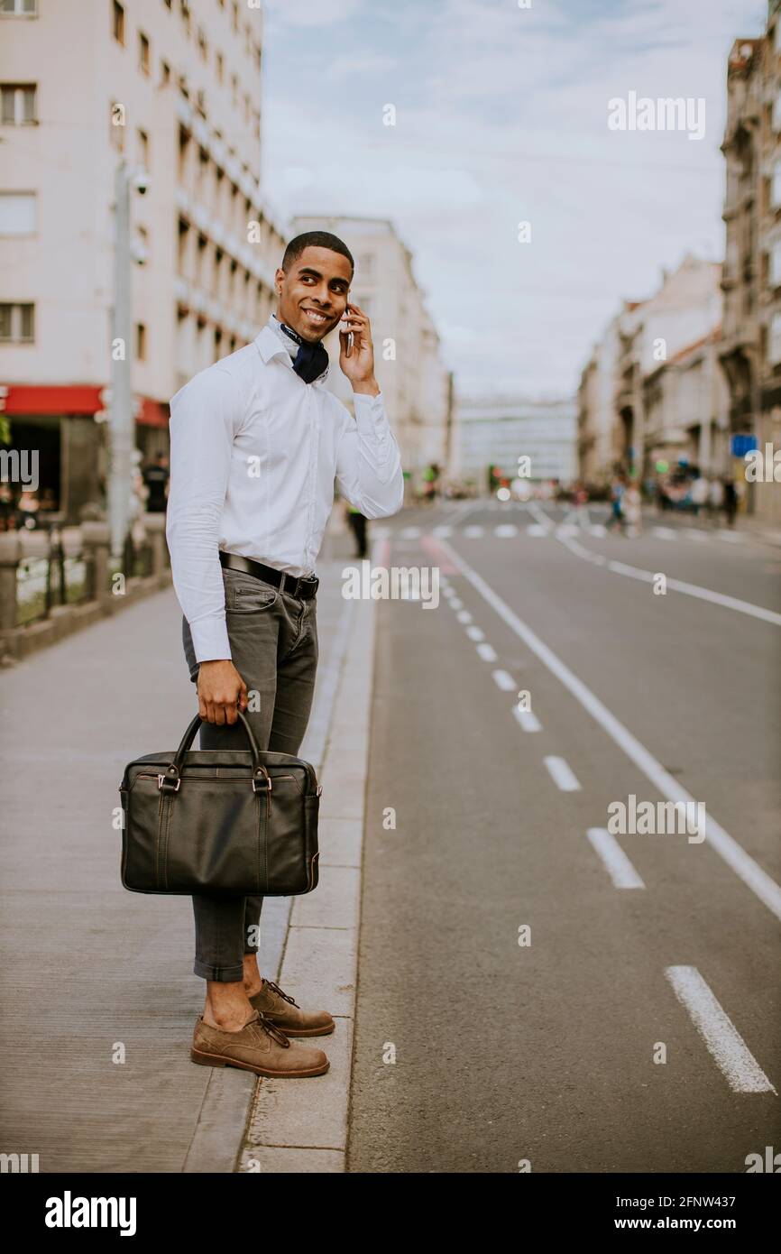 Bel giovane uomo d'affari afro-americano che usa un telefono cellulare mentre attesa di un taxi in attesa di un taxi su una strada Foto Stock