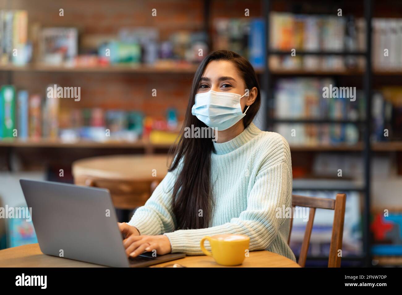 Giovane donna araba in maschera cercando lavoro durante la pandemia di covid-19, usando il laptop in caffetteria, controllando le posizioni vacanti in internet Foto Stock