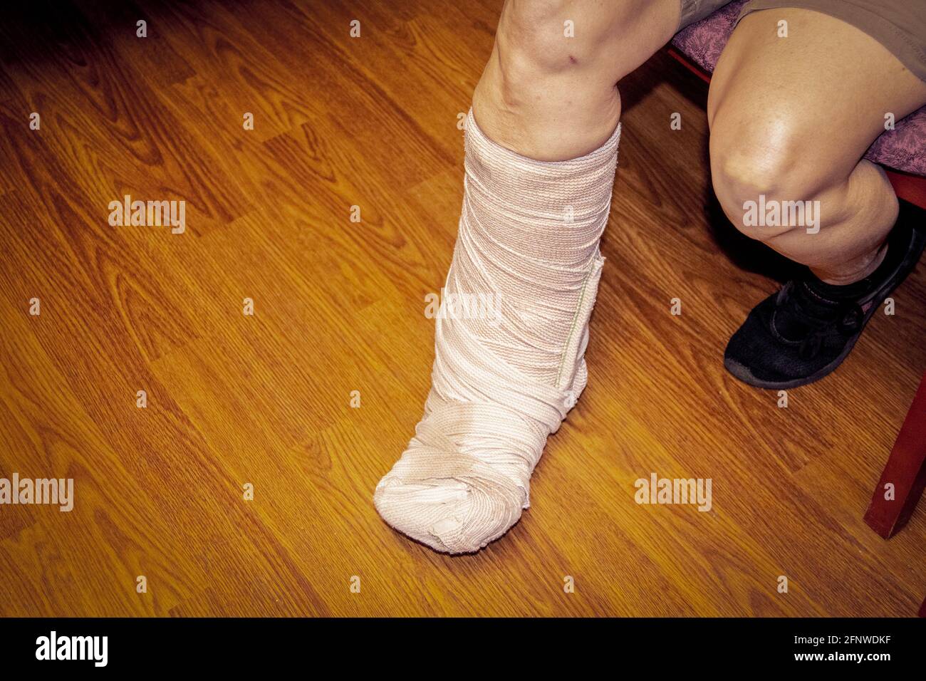 Piede e gamba di donna corto bendato in pantaloncini contro il pavimento di legno come lei si siede su panca o sedia. Foto Stock