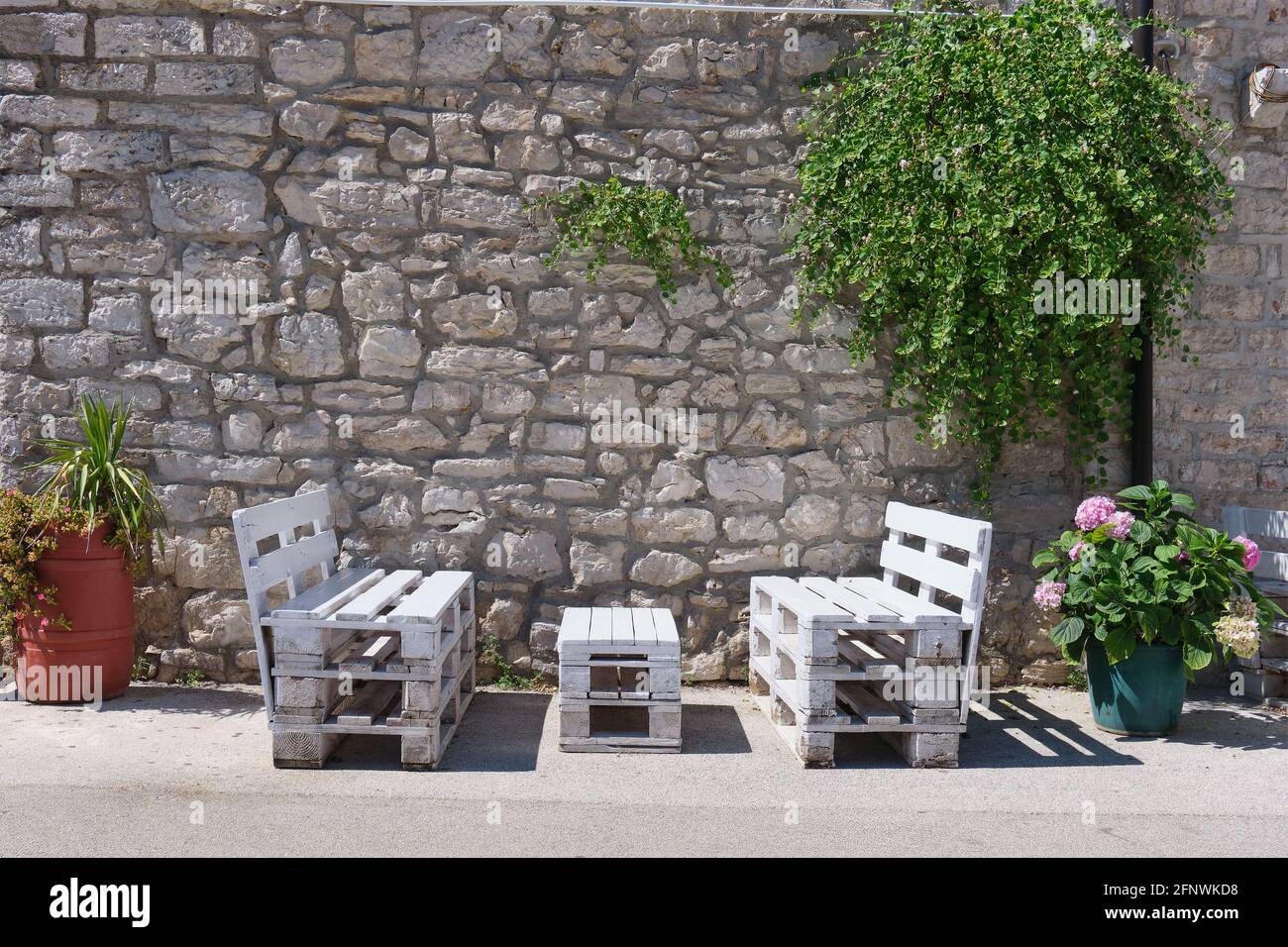 le panchine sono situate sotto il verde di fronte ad un storico muro di pietra tra due vasi di fiori Foto Stock