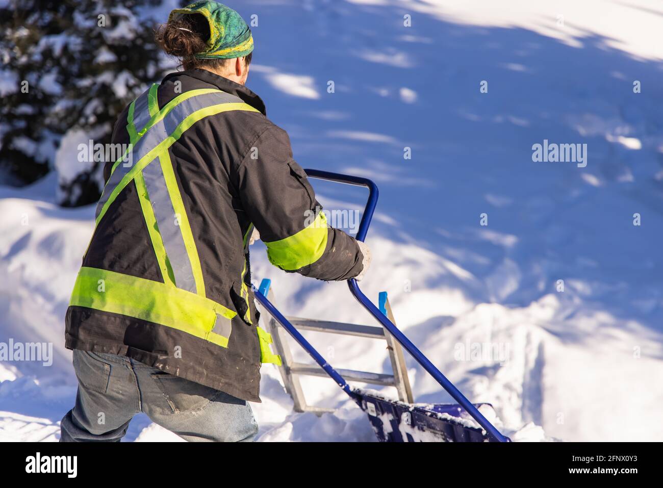 Vista posteriore di un giovane uomo con giacca a visibilità e pala manuale blu che rimuove il ginocchio neve fresca profonda da una casa di campagna durante una soleggiata giornata invernale. Foto Stock