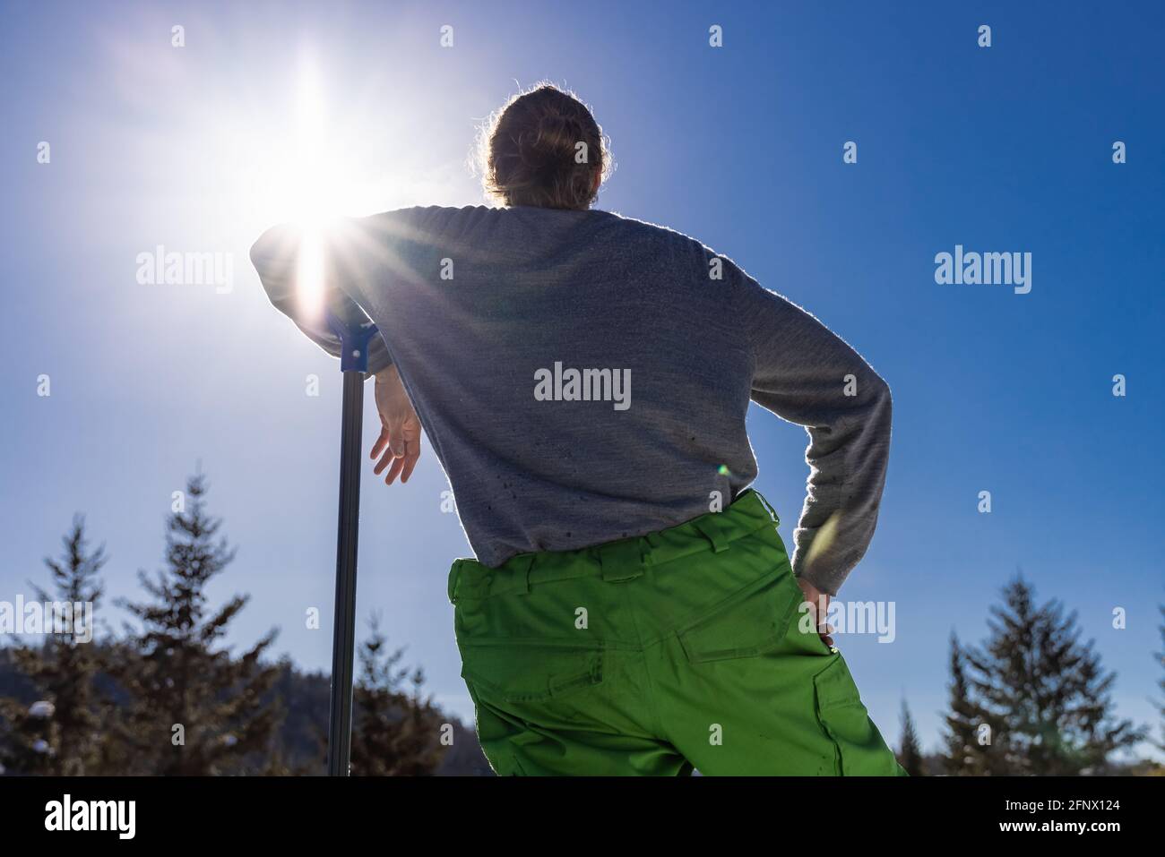 Vista da dietro di un giovane che pende sul manico di una pala da neve blu mentre si fa una pausa durante un lavoro di rimozione della neve in una casa di campagna. Foto Stock