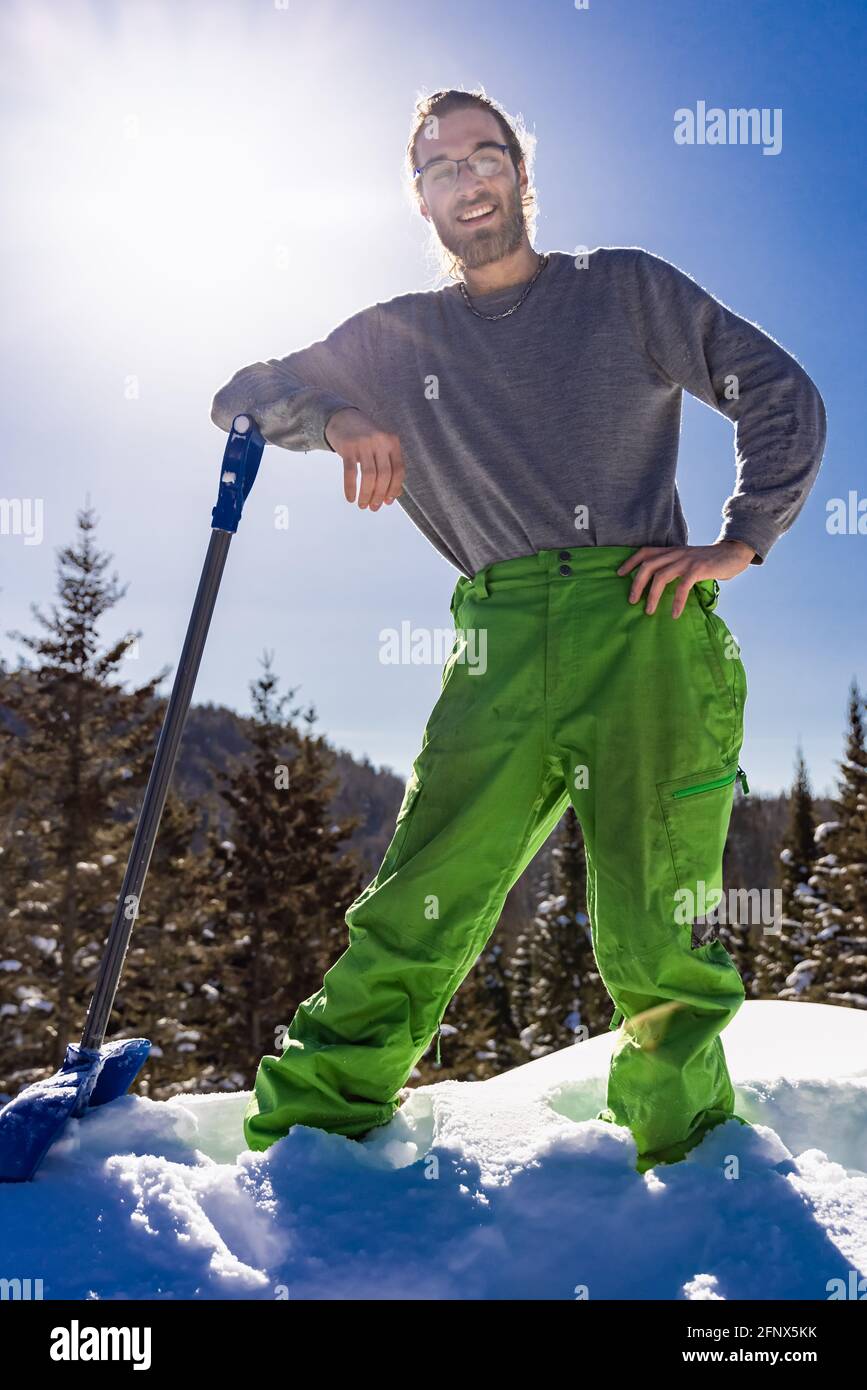 Ritratto a figura intera di un uomo sorridente appoggiato sul manico di una pala da neve blu mentre si fa una pausa durante un lavoro di rimozione della neve in una casa di campagna. Foto Stock