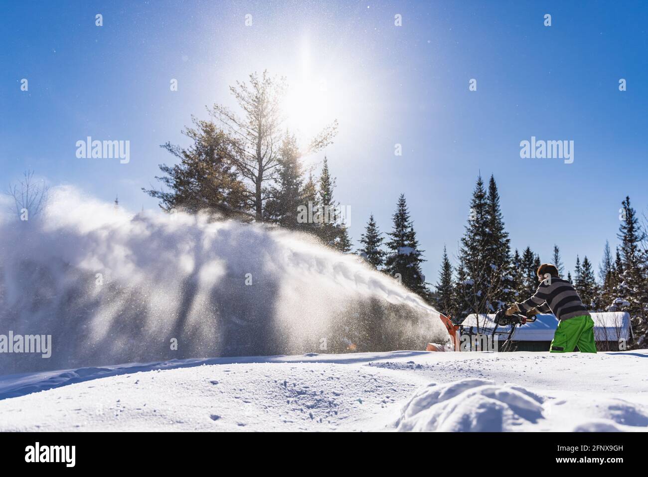 Vista laterale grandangolare di un uomo che crea un'enorme neve scintillante mentre rimuove la neve fresca con uno spazzaneve meccanico in una soleggiata giornata invernale. Foto Stock