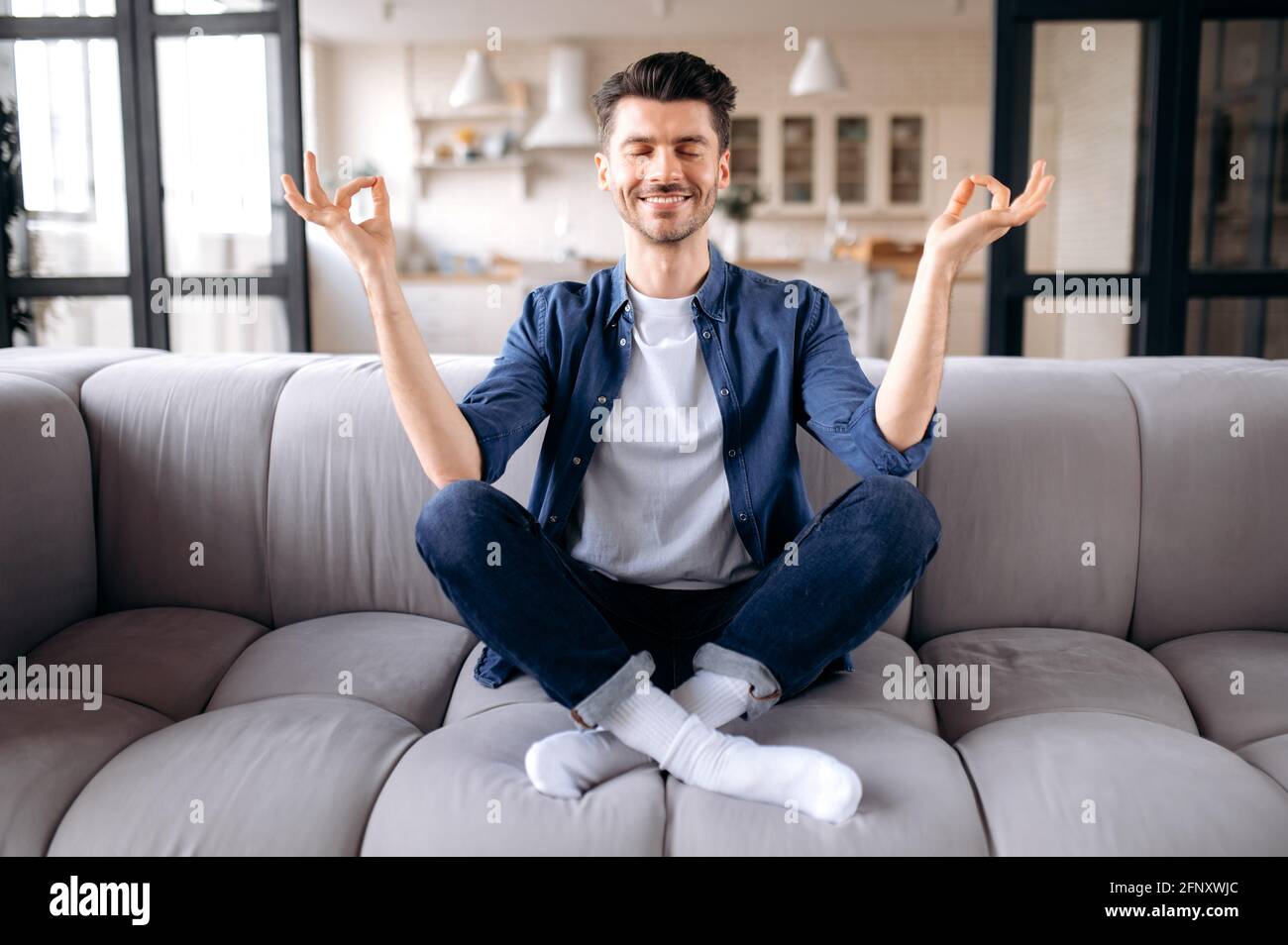 Meditazione, relax, riposo. Caucasica calma soddisfatto ragazzo in abiti casual elegante si siede sul divano nel soggiorno con gli occhi chiusi, medita in posizione lotus, allevia lo stress Foto Stock
