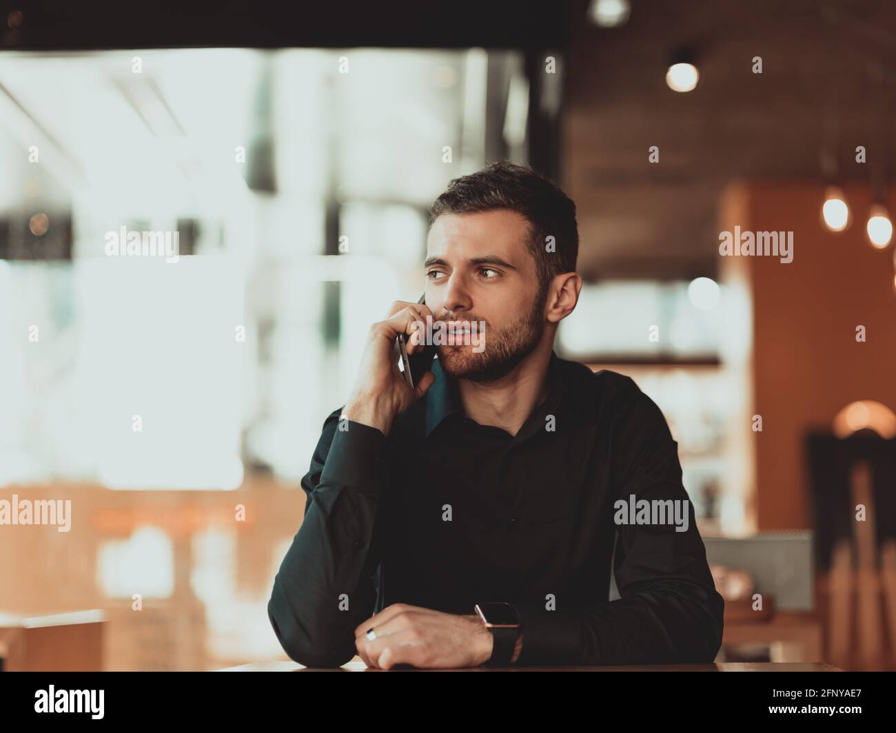 un giovane uomo d'affari in una camicia nera si siede in una caffetteria e usa lo smartphone durante una pausa caffè. Foto Stock