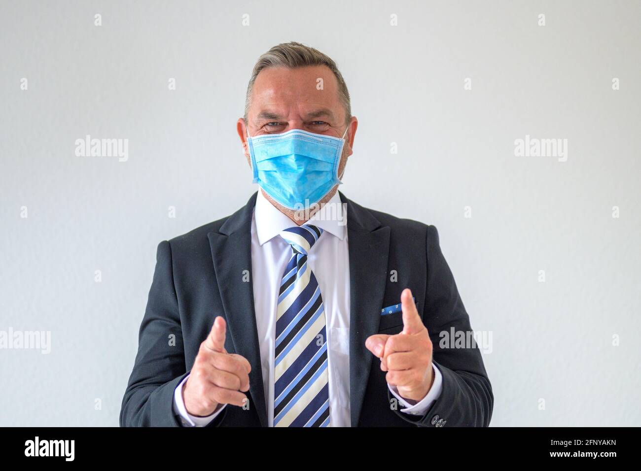 Uomo d'affari felice riuscito in un vestito e legare gesturing con le mani e sorridendo dietro una maschera protettiva durante La pandemia di Covid-19 Foto Stock