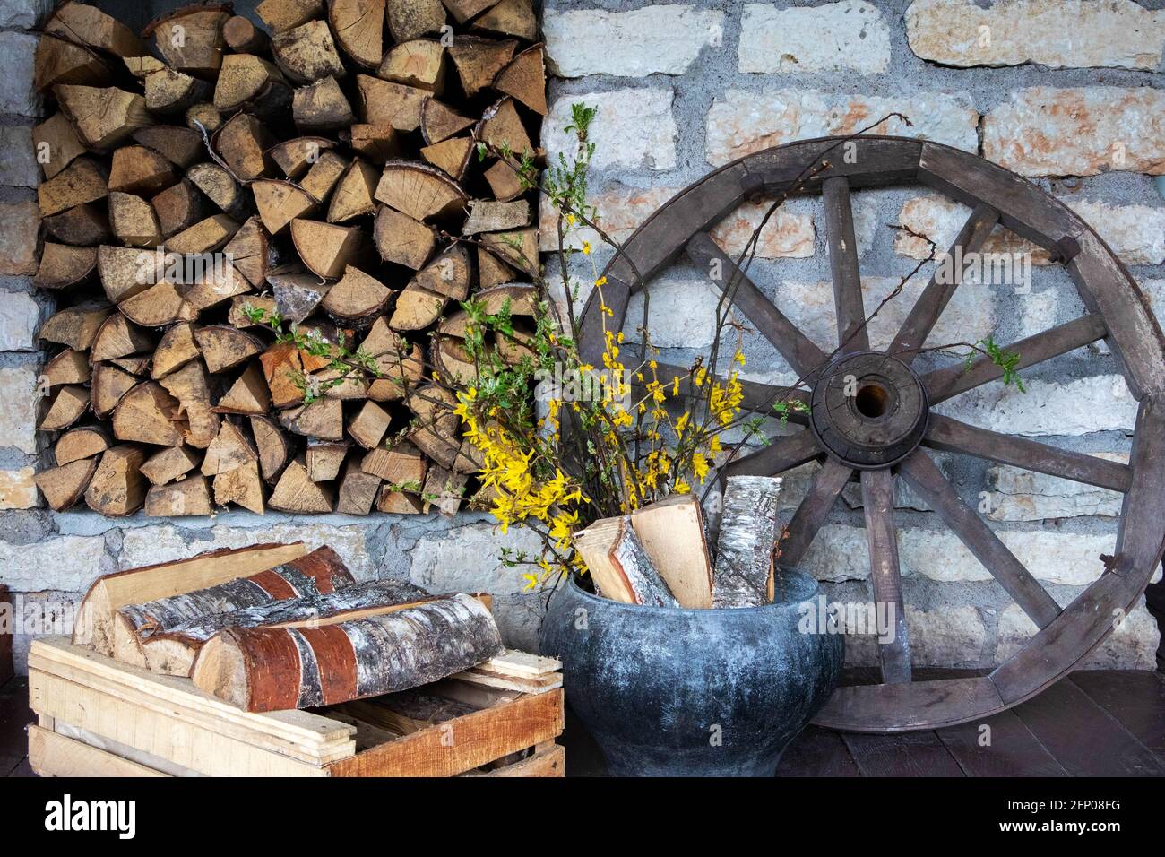 Accessori villaggio, scenario rurale decorativo con vecchia ruota dentata, pila di legna fuoco e vecchia pentola con fiori gialli Foto Stock