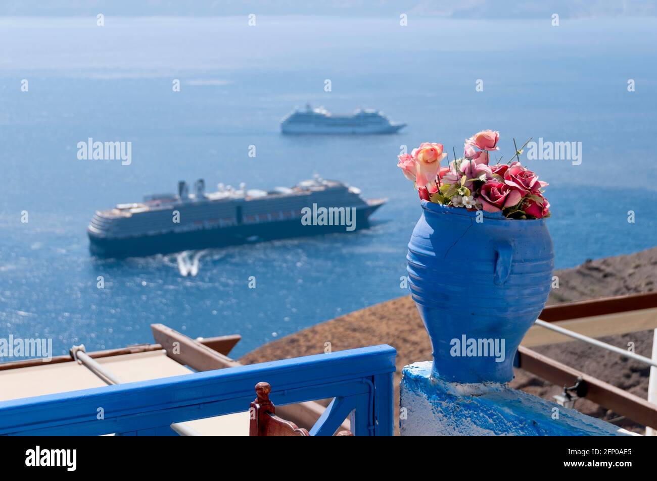 Primo piano di un vaso blu con fiori su una terrazza a Santorini con vista aerea sul mare e sulla caldera. Sullo sfondo le navi da crociera offuscate navigano Foto Stock
