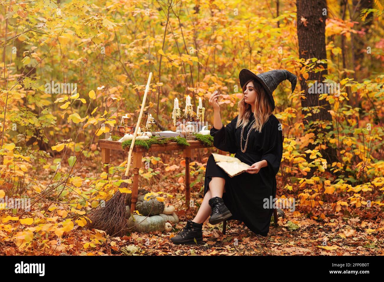 Giovane donna in costume da strega seduta a tavola con magia ingredienti e prendere appunti il giorno d'autunno in foresta Foto Stock