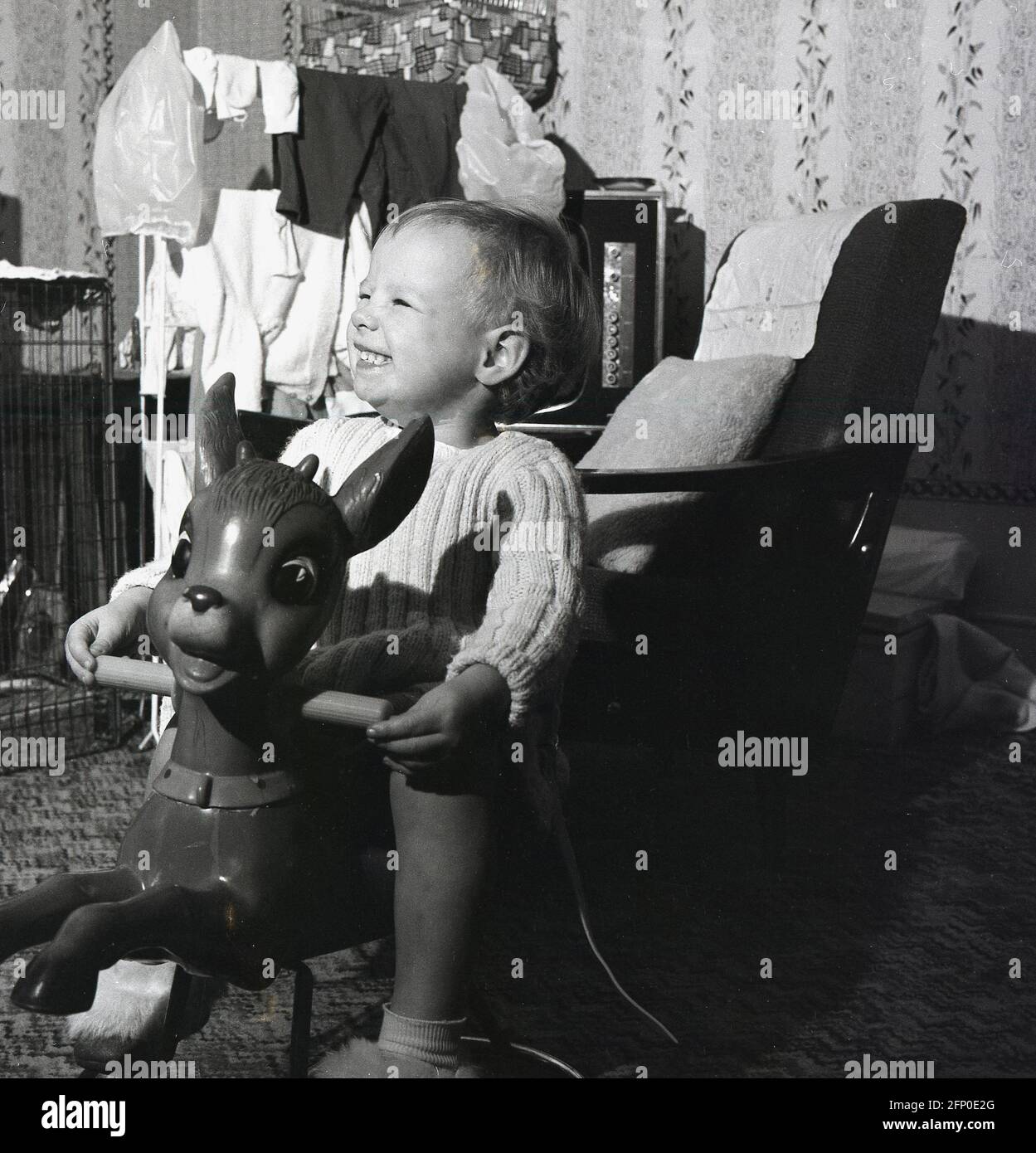 1960s, storico, all'interno di una stanza anteriore, un ragazzino eccitato seduto sul suo cavallo a dondolo giocattolo. Una guardia da fuoco e alcuni vestiti che si asciugano, con una gabbia di uccelli su un televisore anche nella foto. Foto Stock