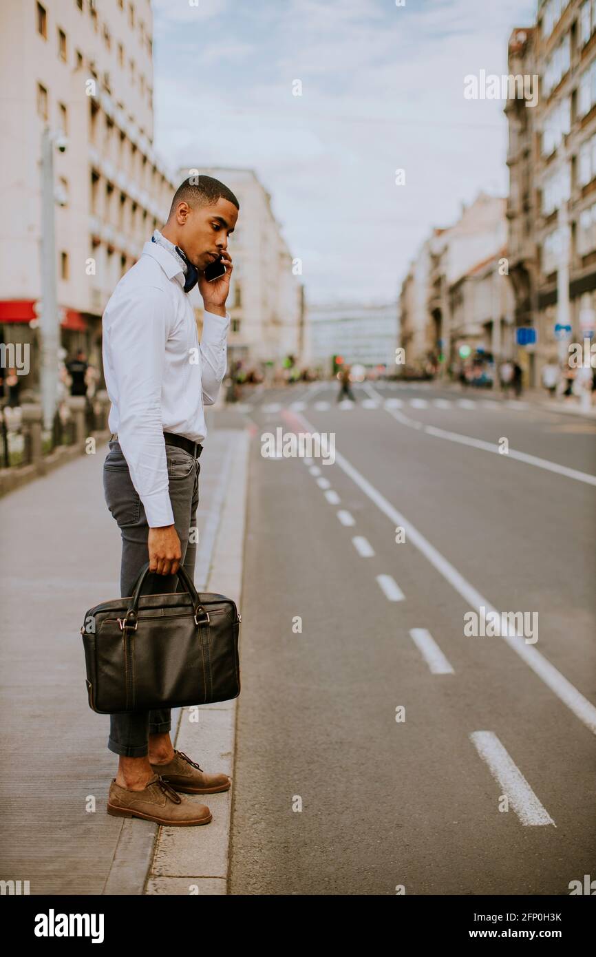 Bel giovane uomo d'affari afro-americano che usa un telefono cellulare mentre attesa di un taxi in attesa di un taxi su una strada Foto Stock