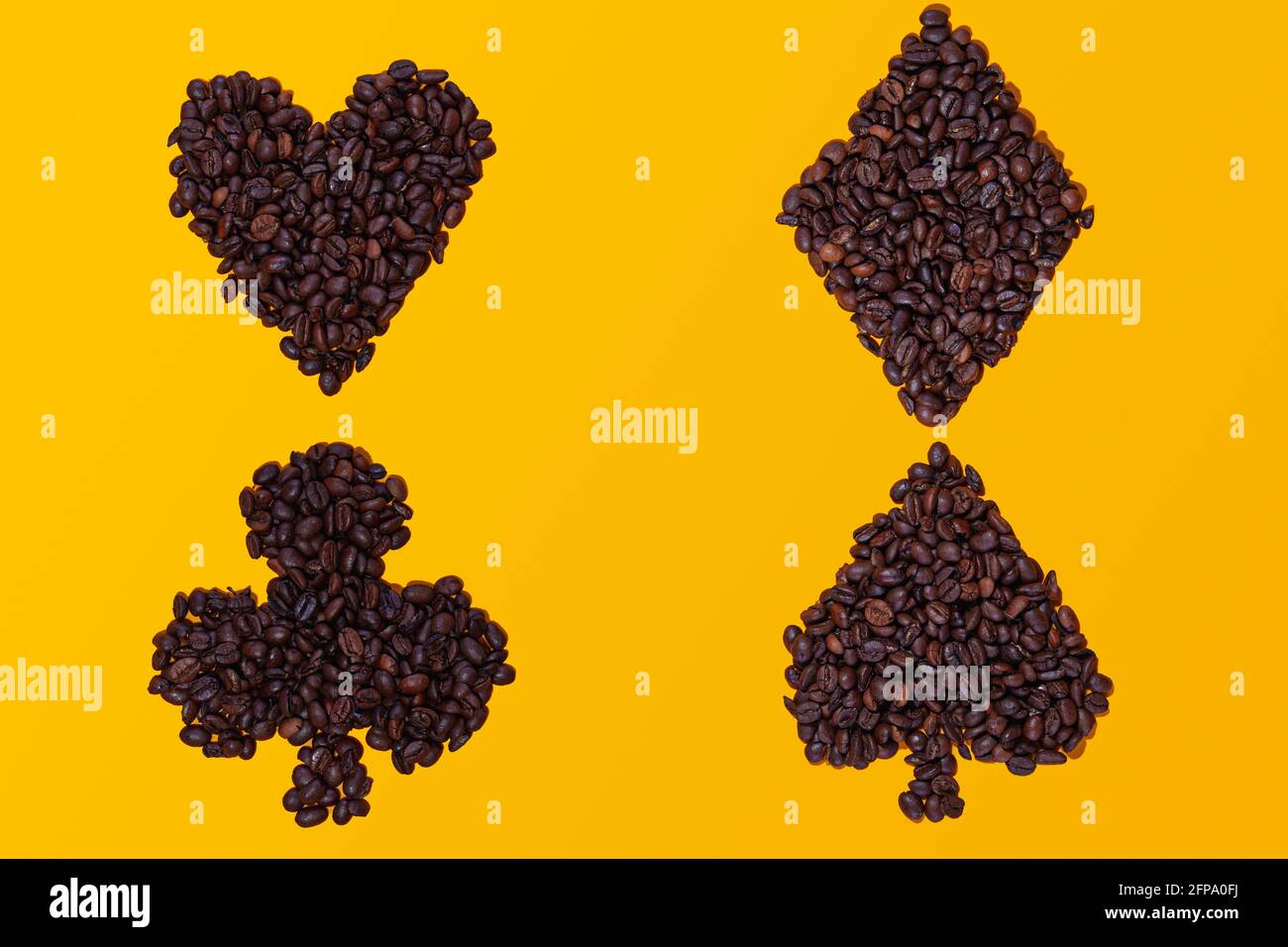 Chicchi di caffè raggruppati nelle forme dei quattro simboli della carta da poker: Cuori, picche, mazze e diamanti. Sono posizionati su uno sfondo giallo. Foto Stock