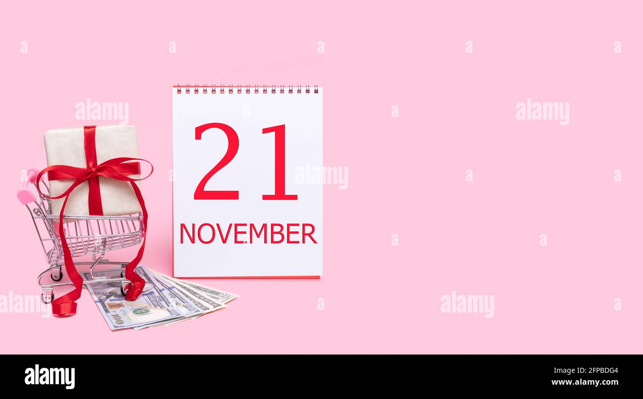 21 novembre. Una confezione regalo in un carrello, dollari e un calendario con la data del 21 novembre su sfondo rosa. Mese di autunno, giorno di Foto Stock