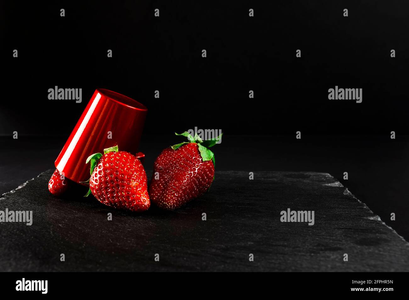 Tre fragole rosse fresche e una tazza rossa su una lavagna. È una vita morta fatta interamente con luce artificiale e sparata in formato orizzontale. Foto Stock