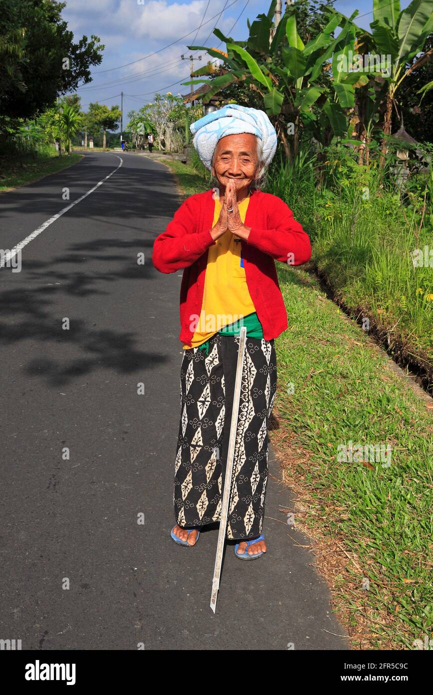 Donna balinese anziana con sarong, cime rosse e gialle, asciugamano avvolto intorno alla testa, in piedi vicino alla strada con le mani in posizione di benedizione della preghiera Foto Stock