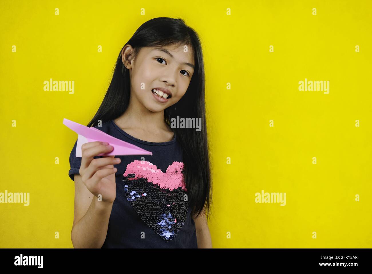 Una giovane ragazza asiatica carina sta giocando con il suo aereo di carta rosa, tenendolo con una mano, puntando e preparandosi al lancio. Sfondo giallo brillante Foto Stock