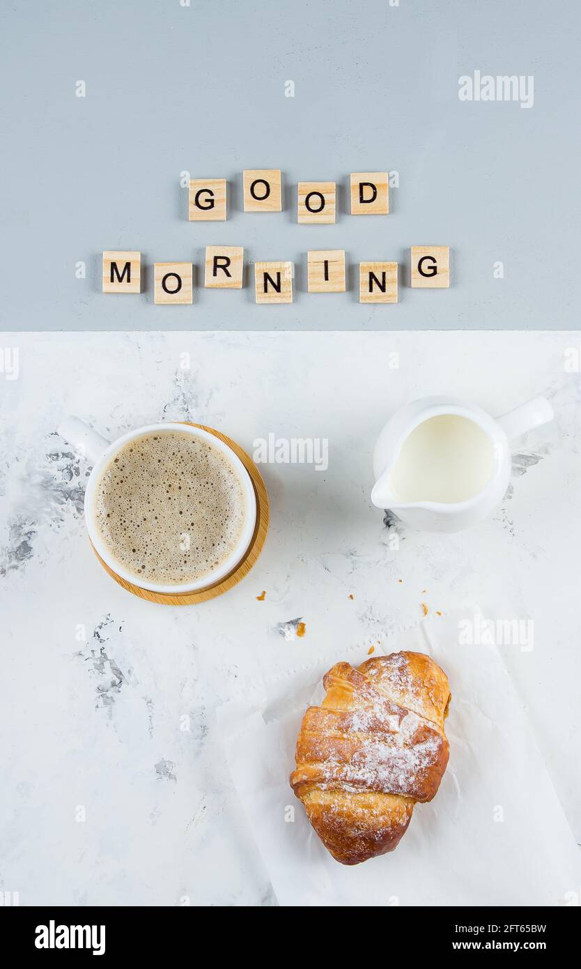Buona colazione del mattino di concetto minimo. Tazza di caffè, croissant e testo Buongiorno su sfondo grigio rosa. Disposizione piatta Foto Stock