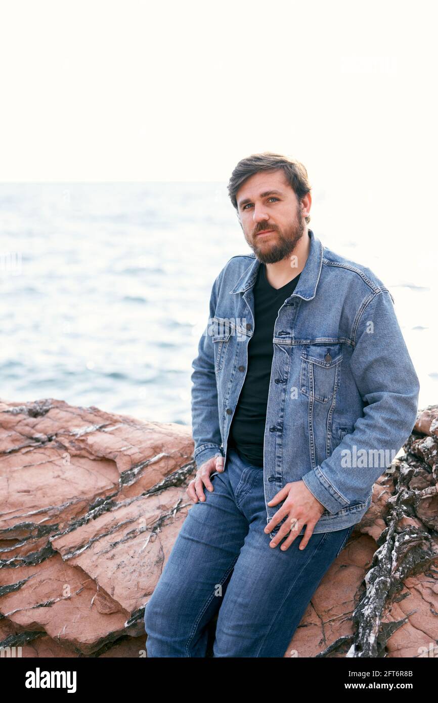 Uomo pensivo in jeans e giacca in denim in posa seduta su pietre sullo sfondo del mare. Vista laterale Foto Stock
