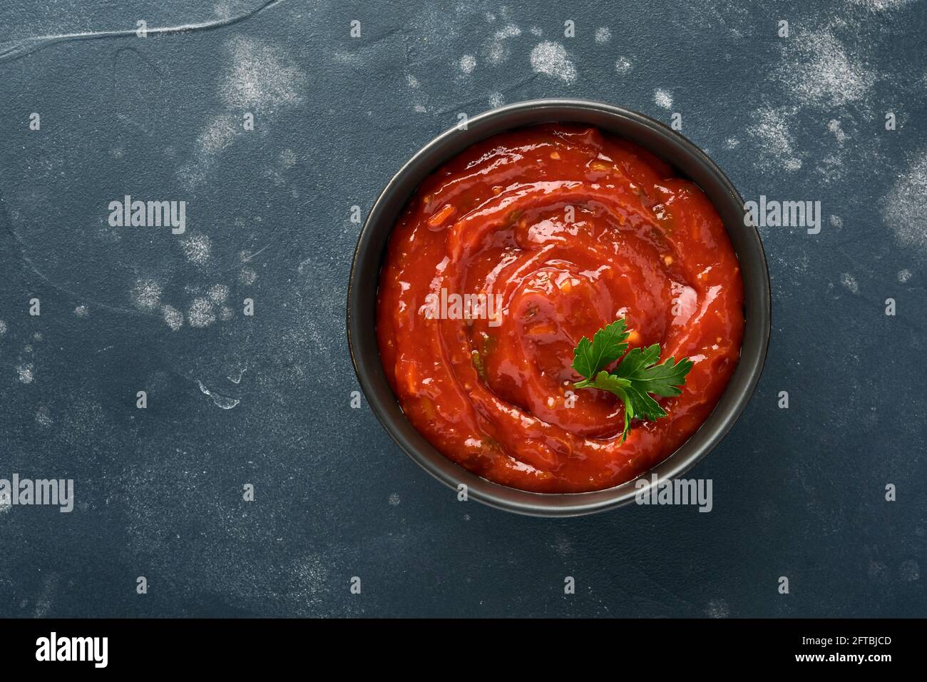 Salsa piccante al ketchup di pomodoro in un recipiente nero con spezie e pomodori  freschi su fondo scuro di ardesia, pietra o cemento. Vista dall'alto con  spazio per la copia Foto stock -