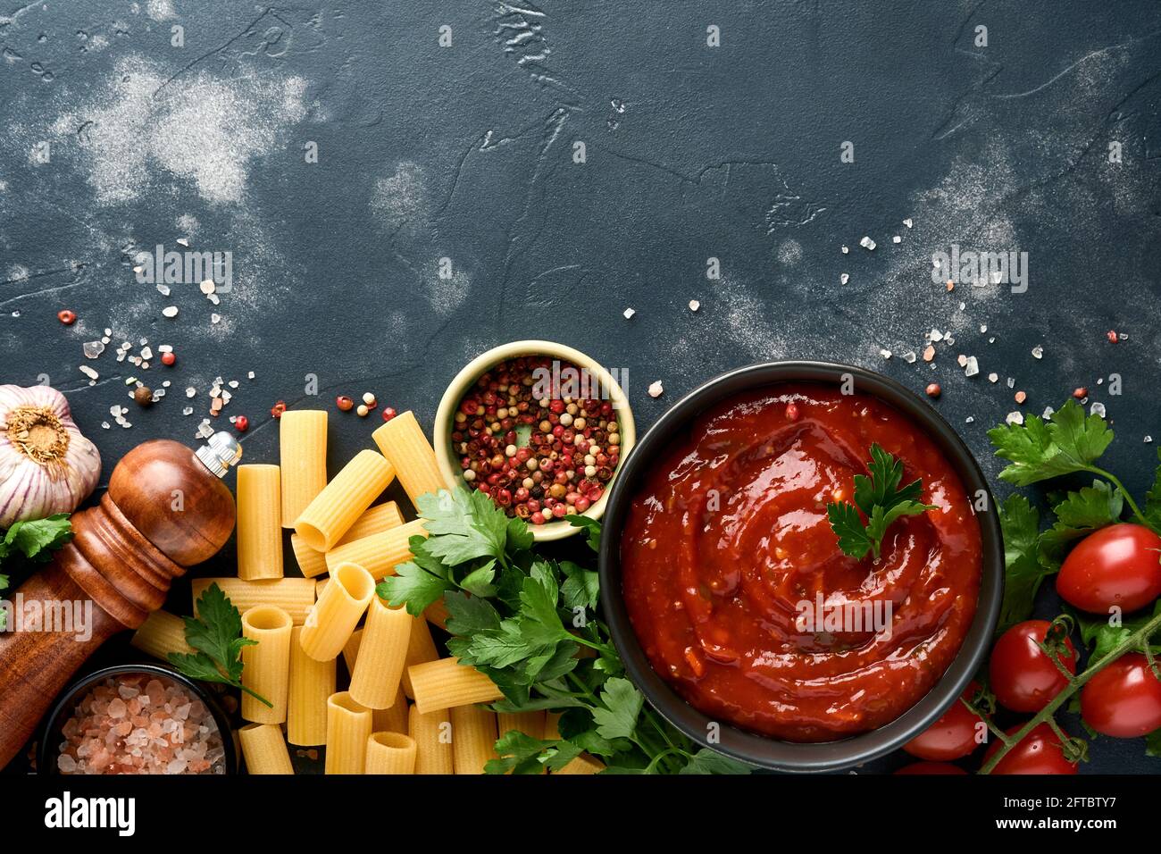 Pasta di fondo. Rigatoni di pasta, salsa di ketchup di pomodoro, olio d'oliva, spezie, prezzemolo, e pomodori freschi su un'ardesia scura, pietra o fondo di calcestruzzo. Foto Stock