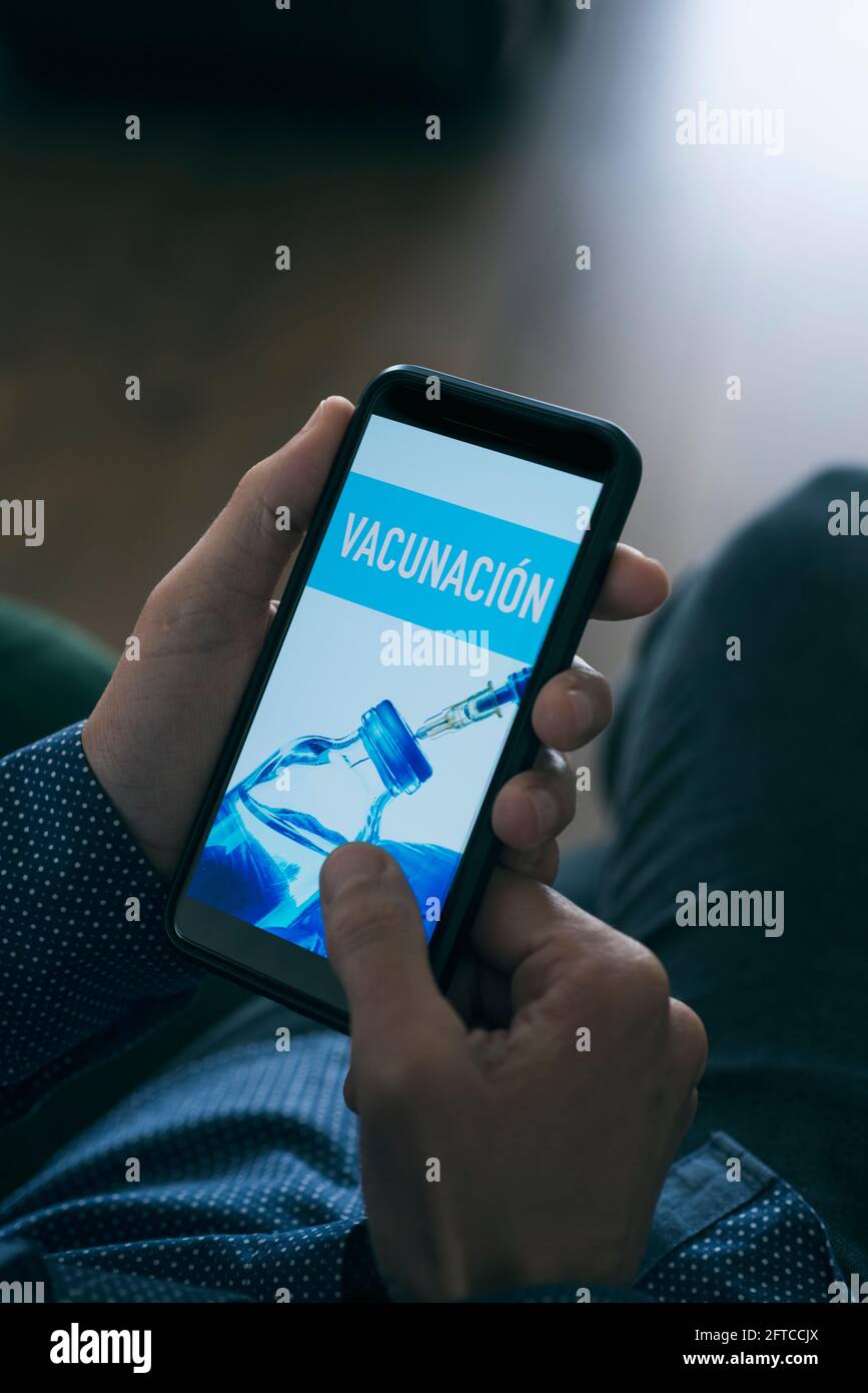 un giovane caucasico, seduto in soggiorno, guarda il suo smartphone, che legge il testo della vaccinazione scritto in spagnolo nel suo schermo Foto Stock