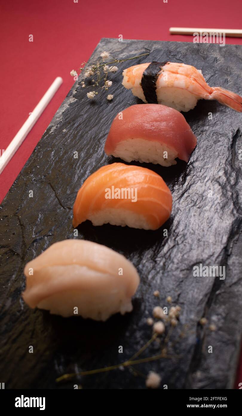 Primo piano di quattro tipi di sushi su un piatto di pietra nera con bastoncini di sushi e due ciuffi di fiori. Foto Stock