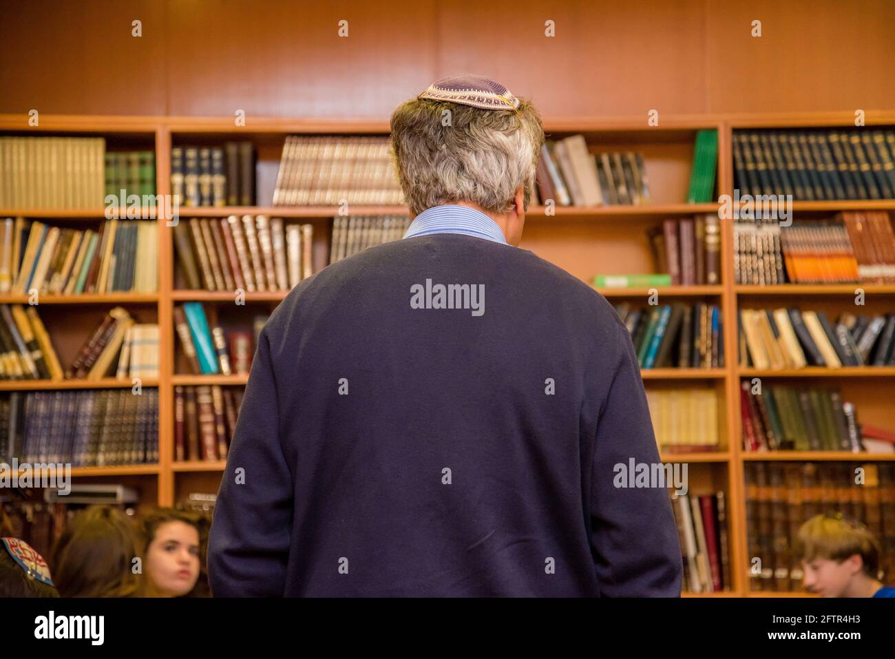 Vecchio ebreo con capelli grigi che indossa un yarmulke dal retro in una biblioteca di scaffali di libri. Foto Stock