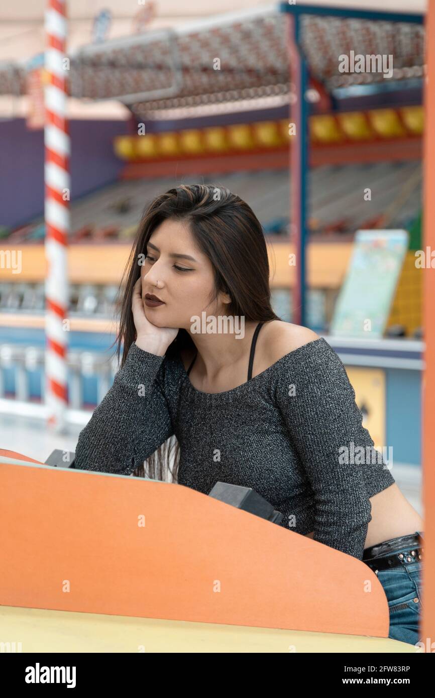 Giovane donna latina che indossa trucco e con capelli neri dritti, seduta triste e annoiato appoggiando la mano al viso, nei giochi di sfondo, stile di vita Foto Stock
