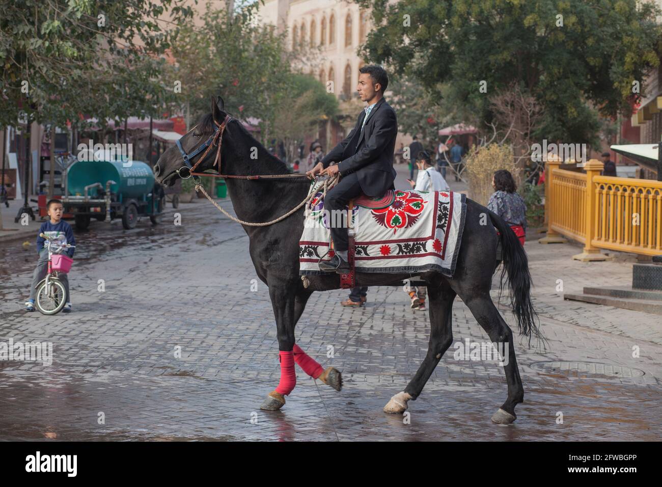 L'uomo sulla cima di un cavallo ornato cammina lungo la strada di Kashgar e un bambino lo guarda Kashgar, Xinkiang, Repubblica popolare di Cina, 2019 Foto Stock