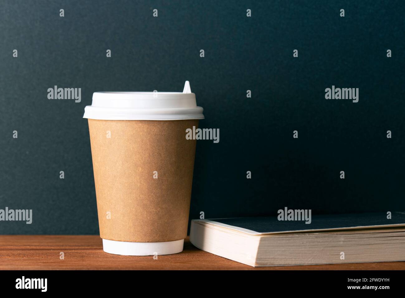 Caffè interno con carta marrone tazza monouso un libro su tavolo di legno di fronte alla parete nera. Composizione minima, spazio per il testo. Interni scuri Foto Stock