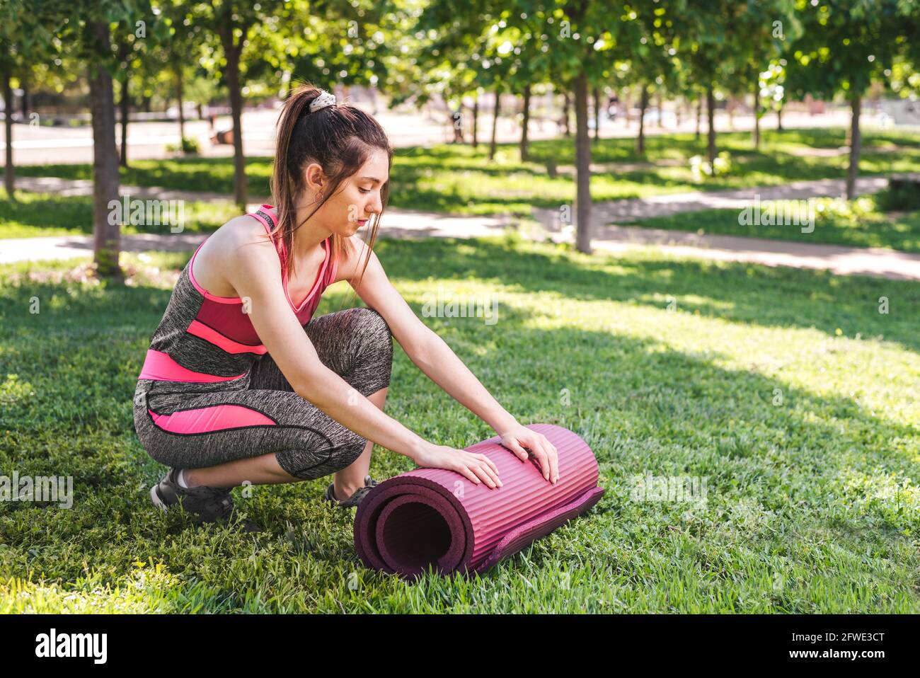 giovane donna caucasica che scopre un opaco yoga sull'erba di un parco verde. Indossa abiti sportivi rosa e grigi Foto Stock