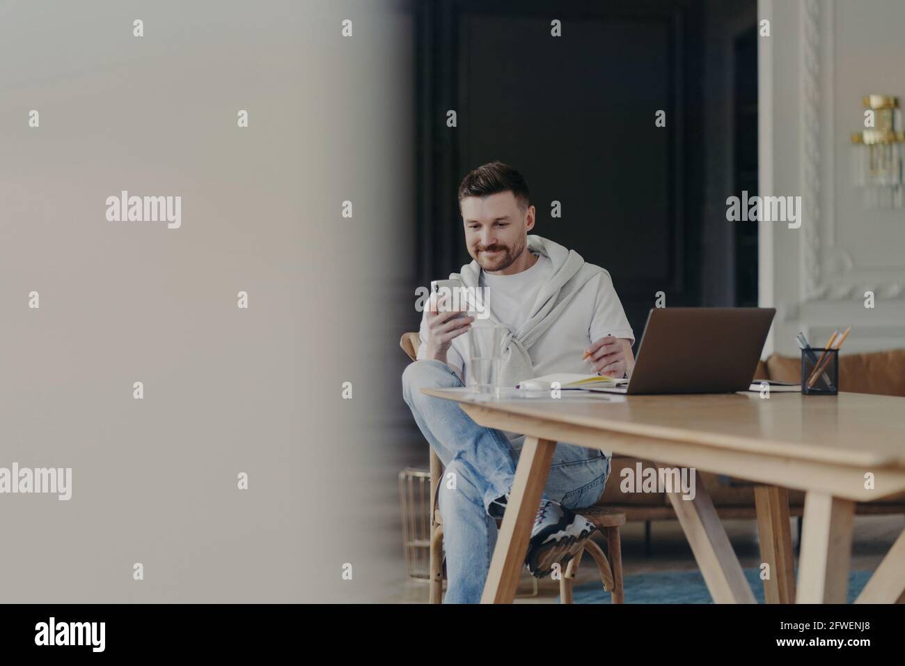 Gioioso giovane imprenditore maschio seduto vicino a tavola di legno e utilizzando smartphone mentre si lavora in remoto su un computer portatile da casa Foto Stock
