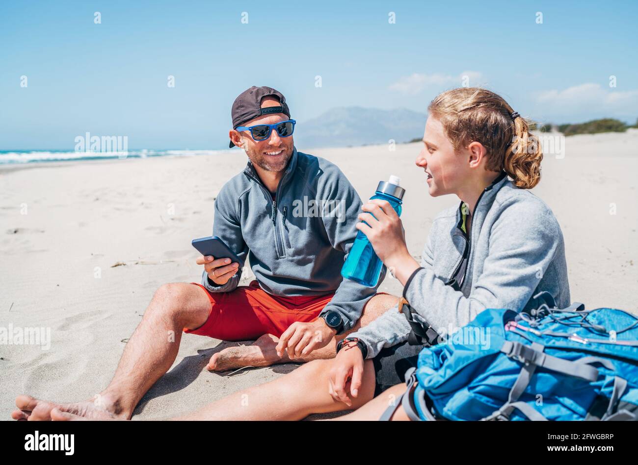 Sorridente Padre in occhiali da sole con smartphone seduto con il figlio adolescente con zaini sulla spiaggia sabbiosa. Ragazzo che beve acqua dalla bottiglia. Acti Foto Stock