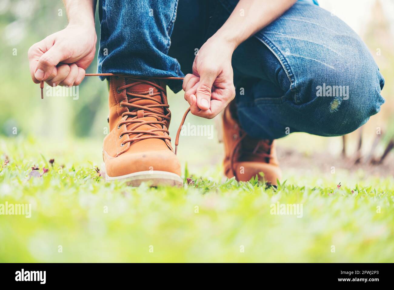 Calzaturiero Concept.Handsome uomo indossare jeans inginocchiati per fare il suo shoelaces. Preparazione prima di andare a festa. Stivali marroni su erba verde. Primo piano MAN han Foto Stock