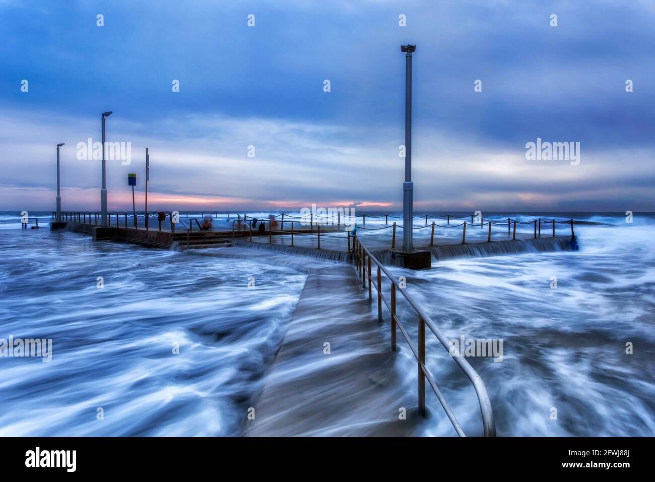 Spiaggia di Mona vale piscina rocciosa ad alta marea tagliata da onde alte durante l'ora di alba blu con nuotatori attivi. Foto Stock